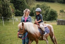 Ein kleiner Junge mit Reithelm sitzt auf einem braun-weißen Pony, während eine blonde Frau neben ihm hergeht und das Pony am Strick führt, im Hintergrund grüne Wiesen rund ums Familienhotel Huber in Südtirol.