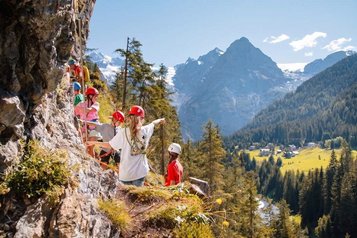 Kinder klettern beim Kinderprogramm mit Helmen an einem Felsen in der Berglandschaft des Hotels Bella Vista in Südtirol.