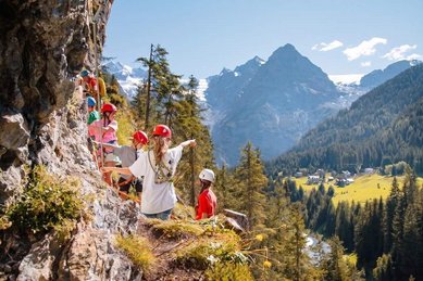 Kinder klettern beim Kinderprogramm mit Helmen an einem Felsen in der Berglandschaft des Hotels Bella Vista in Südtirol.