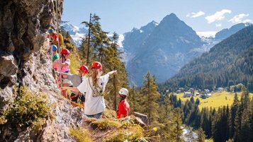 Kinder klettern beim Kinderprogramm mit Helmen an einem Felsen in der Berglandschaft des Hotels Bella Vista in Südtirol.