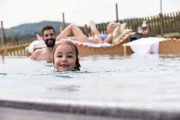 Kind schwimmt lachend im Außenpool, während Erwachsene im Hintergrund auf Liegen entspannen.