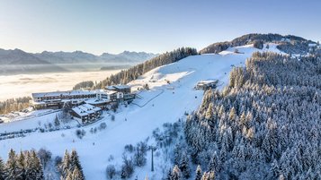 Verschneites Skigebiet in den Alpen mit Hotelanlage am Hang, Skipisten, Sessellift und dichtem Nadelwald im Morgenlicht über dem Nebel im Tal