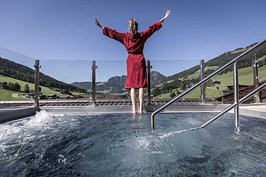 Frau steht im Bademantel am Outdoor-Pool des Familienhotels Galtenberg Family & Wellness Resort und streckt die Arme in die Höhe.