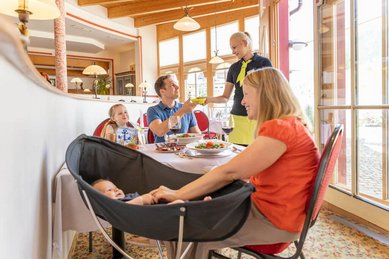 Familie mit Baby und Kleinkind genießt das Essen im Restaurant des Familienhotel Kaiserhof, während eine Kellnerin Getränke serviert.