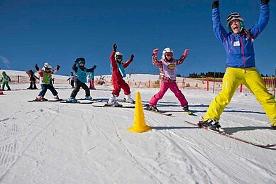 Die Kinder der Kinderskischule fahren ihrer Skilehrerin hinterher und heben dabei ihre Arme in die Höhe. Die Skischule befindet sich ganz in der Nähe des im Familienhotels Feldberger Hof im Hochschwarzwald.