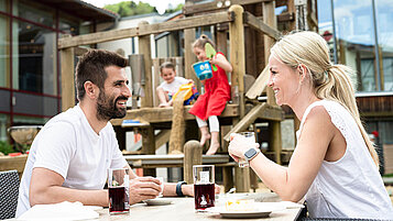 Familienhotels mit Kinderspielplätzen für ganz viel Spaß. Eltern genießen etwas Zeit zu zweit. Im Hintergrund spielen die Töchter auf einem Klettergerüst.