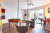 Geräumiger Freizeitraum im Familienhotel Deichkrone mit einem Tischfußball, bequemen Sitzgelegenheiten, einem Projektor und Zugang zu einer Terrasse, ideal für Teenager und Familienunterhaltung.