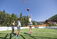 Kinder spielen auf dem Spielplatz des Familienhotels Galtenberg Family & Wellness Resort Basketball bei gutem Sommer Wetter.