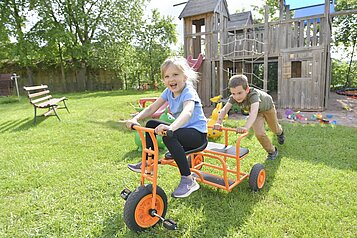Lachendes Mädchen sitzt auf einem Dreirad und wird von einem Jungen angeschoben. Im Hintergrund ist der Außenspielplatz des Familienhotels Rhön Feeling zu sehen.