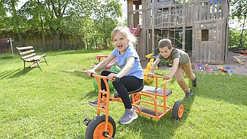 Lachendes Mädchen sitzt auf einem Dreirad und wird von einem Jungen angeschoben. Im Hintergrund ist der Außenspielplatz des Familienhotels Rhön Feeling zu sehen.