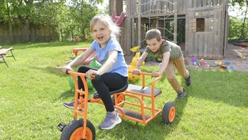 Lachendes Mädchen sitzt auf einem Dreirad und wird von einem Jungen angeschoben. Im Hintergrund ist der Außenspielplatz des Familienhotels Rhön Feeling zu sehen.
