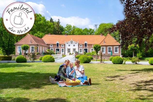 Familie beim Picknick auf einer Wiese vor einem historischen Gutshaus im Hotel Gut Landegge im Emsland mit rotem Ziegeldach, umgeben von Bäumen und Gartenanlage, mit „Familotel der Woche“-Siegel.