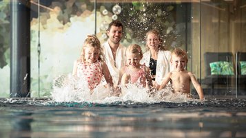 Familie mit drei Kindern planscht fröhlich im Indoor-Pool eines Familienhotels und spritzt mit Wasser.