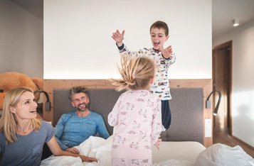 Vierköpfige Familie tobt auf dem Bett im Familienzimmer des Familienhotels Alpenhof Dolomit Family in Südtirol.
