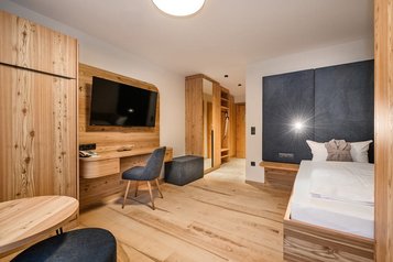 Modernes Familienhotelzimmer mit viel Holz, gemütlichem Bett, großem Fernseher, Schreibtisch und offenem Eingangsbereich für ein warmes Wohngefühl.