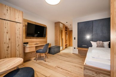Modernes Familienhotelzimmer mit viel Holz, gemütlichem Bett, großem Fernseher, Schreibtisch und offenem Eingangsbereich für ein warmes Wohngefühl.