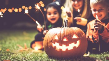 Ein für Halloween beleuchteter und geschnitzter Kürbis, im Hintergrund sitzen drei Kinder 