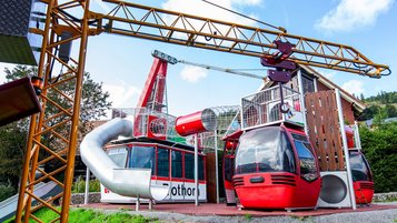 Spielplatz aus umgebauten roten Gondelkabinen mit Rutschen, Kletterelementen, Gittern und einem gelben Kranarm, eingebettet in eine grüne Landschaft unter blauem Himmel.