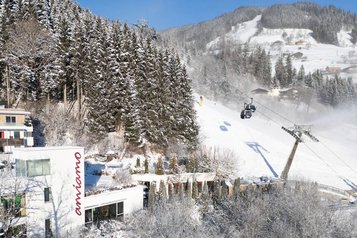 Winteransicht des Familienhotels Amiamo direkt an der Skipiste mit verschneiten Bäumen und Seilbahn im Hintergrund.