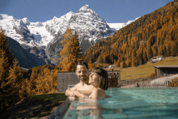 Zwei Personen entspannen im Infinitypool des Hotels Bella Vista in Südtirol mit Blick auf herbstlich gefärbte Bäume und schneebedeckte Berge.
