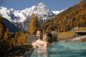 Zwei Personen entspannen im Infinitypool des Hotels Bella Vista in Südtirol mit Blick auf herbstlich gefärbte Bäume und schneebedeckte Berge.