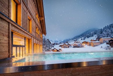 Beheizter Außenpool des Hotels Gorfion in Liechtenstein vor verschneitem Bergdorf am Abend, während Schneeflocken fallen und warme Lichter aus den Häusern leuchten.