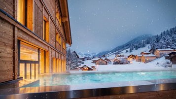 Beheizter Außenpool des Hotels Gorfion in Liechtenstein vor verschneitem Bergdorf am Abend, während Schneeflocken fallen und warme Lichter aus den Häusern leuchten.
