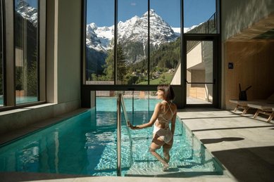 Eine Frau steht im Hallenbad vor der Glasschleuse zum beheizten Outdoor-Infinity-Panoramapool, daneben ein Freiluft-Whirlpool, im Hotel Bella Vista in Südtirol.