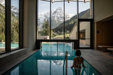 Eine Frau steht im Hallenbad vor der Glasschleuse zum beheizten Outdoor-Infinity-Panoramapool, daneben ein Freiluft-Whirlpool, im Hotel Bella Vista in Südtirol.
