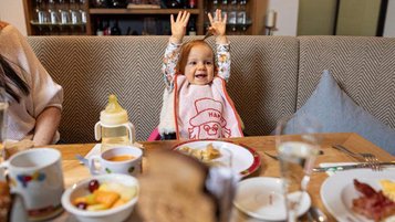Kleinkind sitzt jauchzend am Familientisch im Landgut Furtherwirt in Tirol. Mit Kids All-Inclusive sind eure Kleinen bestens versorgt. 