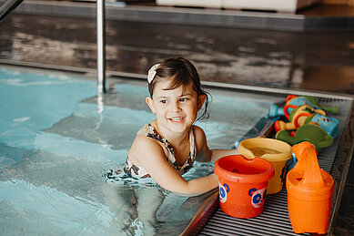 Kleines Mädchen spielt mit Eimer und Gießkanne am Beckenrand des Indoor-Schwimmbeckens im Familienhotel Huber in Südtirol.