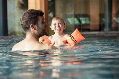 Vater und Kind lachen gemeinsam im Schwimmbad, das Kind trägt orange Schwimmflügel.