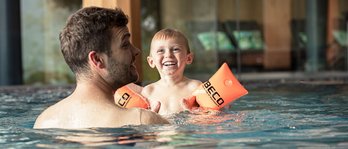 Vater und Kind lachen gemeinsam im Schwimmbad, das Kind trägt orange Schwimmflügel.