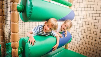 Zwei Kinder haben Spaß im Indoor-Spielbereich des Landguts Furtherwirt und klettern lachend durch weiche, grüne Hindernisse