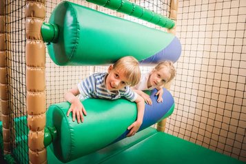 Zwei Kinder haben Spaß im Indoor-Spielbereich des Landguts Furtherwirt und klettern lachend durch weiche, grüne Hindernisse