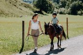 Eine Erwachsene begleitet ein Kind auf einem Pony auf einem Weg im Außenbereich des Familienhotels Oberkarteis im Salzburger Land.
