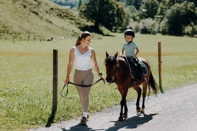 Eine Erwachsene begleitet ein Kind auf einem Pony auf einem Weg im Außenbereich des Familienhotels Oberkarteis im Salzburger Land.