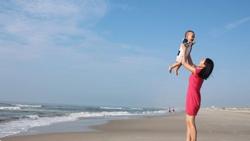 Mutter steht mit ihrem Baby am Strand und hebt es in die Höhe.