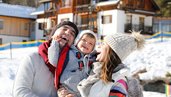 Familie mit Baby lacht gemeinsam im verschneiten Außenbereich des Familienhotels Ramsi in Kärnetn