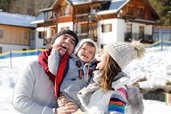 Familie mit Baby lacht gemeinsam im verschneiten Außenbereich des Familienhotels Ramsi in Kärnetn