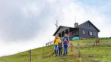 Rhön Wandern: Familie wandert auf einem Berg. Im Hintergrund eine typische Berghütte