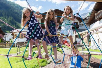 Drei Kinder spielen im Sommer auf dem Outdoor-Spielplatz des Familienhotels Alpenhotel Kindl in Tirol. Auf dem Outdoor Spielplatz hat man einen herrlichen Blick auf die umliegenden Berge.