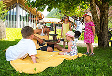 Ein Kinderbetreuer spielt Gitarre und die Kinder hören aufmerksam zu im Garten des Familienhotels Spa- & Familien-Resort Krone im Allgäu.