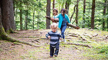 Drei Kinder laufen durch den Wald uns spielen mit Ästen im Familienurlaub in der Steiermark.