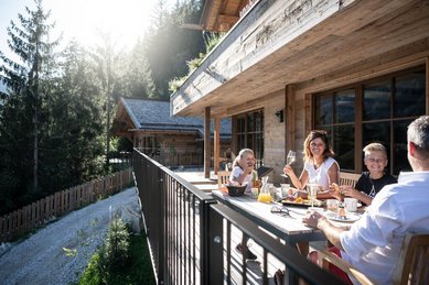 Familie genießt gemeinsam ein Frühstück an einem Holztisch auf der sonnigen Terrasse eines Chalets im Alpura Retreat inmitten der Natur.