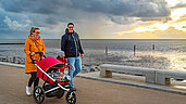 Eine glückliche Familie genießt einen entspannten Spaziergang auf dem Deich an der Nordsee, wobei die Eltern einen Kinderwagen schieben und ein malerischer Sonnenuntergang über dem ruhigen Meer den Hintergrund bildet.