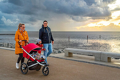 Eine glückliche Familie genießt einen entspannten Spaziergang auf dem Deich an der Nordsee, wobei die Eltern einen Kinderwagen schieben und ein malerischer Sonnenuntergang über dem ruhigen Meer den Hintergrund bildet.