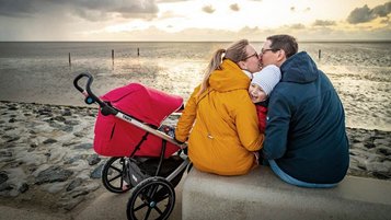Eine glückliche Familie genießt einen entspannten Spaziergang auf dem Deich an der Nordsee, wobei die Eltern einen Kinderwagen schieben und ein malerischer Sonnenuntergang über dem ruhigen Meer den Hintergrund bildet.