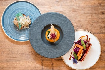 Drei unterschiedlich gestaltete Teller mit kreativ angerichteten Speisen, darunter Gemüse, Fleisch und ein Gericht mit Sprossen, serviert auf einem Holztisch im Familienhotel Huber in Südtirol.