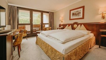 Das Deluxe-Schlafzimmer Familiennest Süd im Hotel Sonngastein im Salzburger Land zeigt ein Doppelbett, einen Schreibtisch und einen Balkon mit Blick nach draußen.
